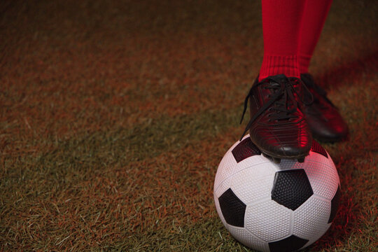 Soccer ball is being held under black cleat, red knee-high sock on trimmed grass, copy space