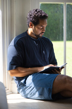 Mid adult African American man sitting on window seat at home reading book with sunlight streaming