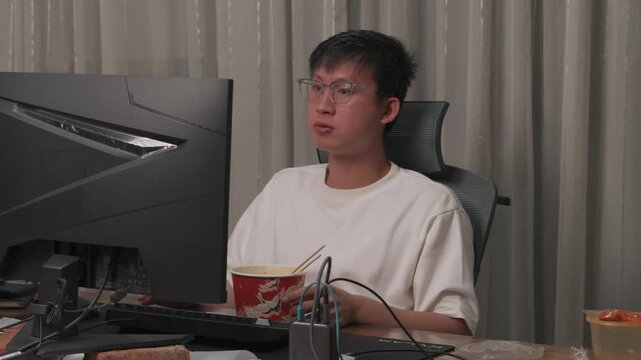 Young man sits at desk, drinking from cup. He eats noodles while looking at computer screen. Caffeine buzz and focused work energy. Relaxed atmosphere at home office space. Good for depicting work
