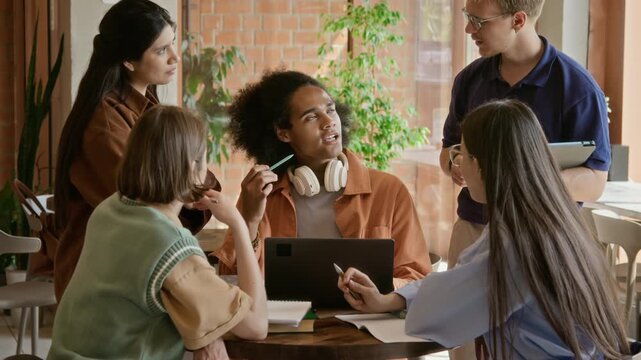 Multiethnic group of young adult students collaborating around table with laptop and notebooks in library. Footage is suitable for university marketing and teamwork ads