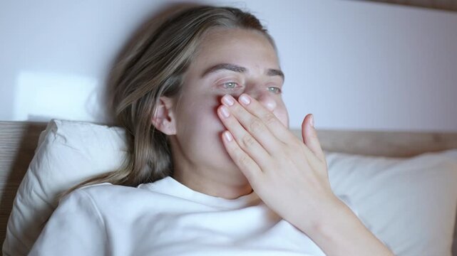Caucasian woman with long hair sits on bed, thoughtfully gazing at a screen, with soft lighting illuminating her face and pillows in a cozy bedroom setting