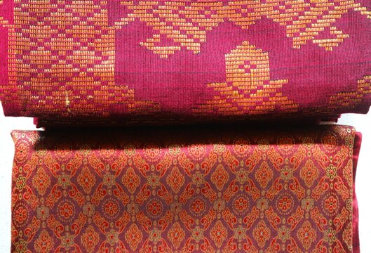 Traditional Indonesian Songket fabric, characterized by shimmering gold or silver thread motifs woven against maroon and pink backgrounds.