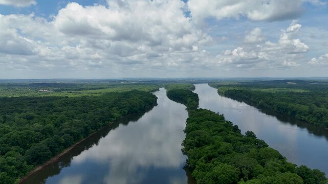 Aerial drone footage over the Potomac River at Algonkian Regional Park in Sterling, Virginia. The river flows around lush green forest islands under a blue sky with fluffy white cloud reflections.