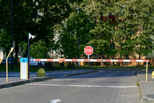 Automatic access control boom barrier arm installed with cctv camera and red stop sign at residential driveway entrance under green trees background. Security boom gate restricting car traffic