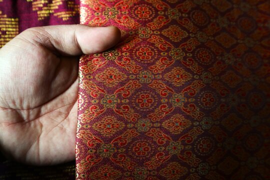 A piece of traditional songket fabric featuring a maroon background and intricate gold thread (zari) weaving, forming a dense, luxurious floral-geometric pattern.
