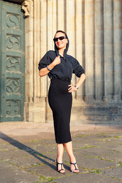 Portrait young adult elegant woman black dress and sunglasses smiles confidently in golden sunlight warm sunset backlit light against historic cathedral building. Female person walking city street