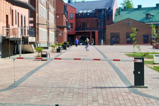 Barrier gate blocking brick paved pedestrian street in renovated urban district while several people walk along walkway. Lowered boom barrier closing wide cobblestone walkway in historic city quarter