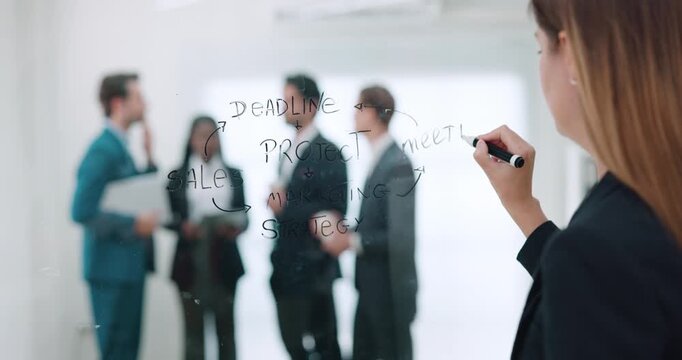 Caucasian woman writes Meeting on glass board while multicultural colleagues talk in background. Businesswoman marks planning notes building workflow map