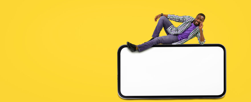Smiling African American Man Relaxing While Sitting On Top Of Empty Smartphone Screen, Holding Phone At Home With Joyful Expression, Engaging In Digital Communication. Technology Concept.