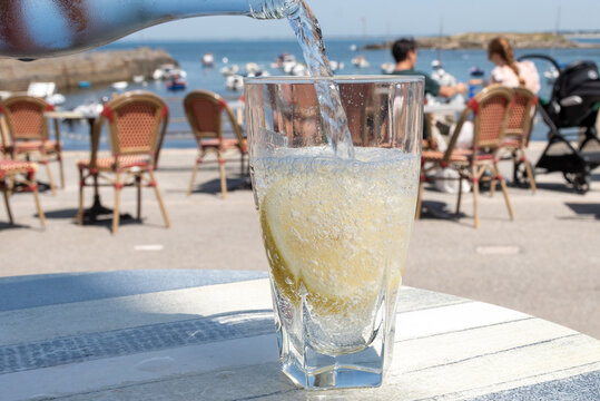 Mineral water pouring into clear glass with fresh lemon slice in French cafe on seaside, hot weather conditions