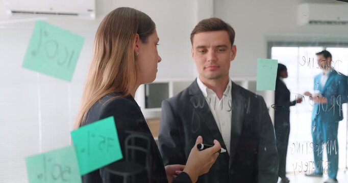 Caucasian businesswoman writes note on glass board by serious man colleague. Woman marks planning idea continuing project talk during focused office brainstorm