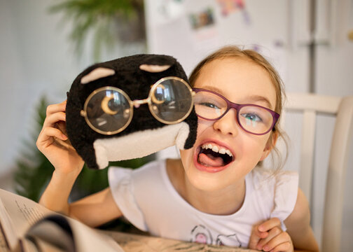 Playful girl with glasses laughs with a plush toy wearing funny spectacles at home. Happy child poses beside an open book, creating a joyful, humorous childhood and reading scene.