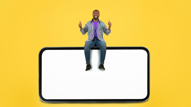 Happy African American Man Sitting On Top Of Smartphone With Blank Screen, Giving Thumbs Up Gesture At Studio. Positive Vibe In Business Environment, Promoting New App Or Website Concept.