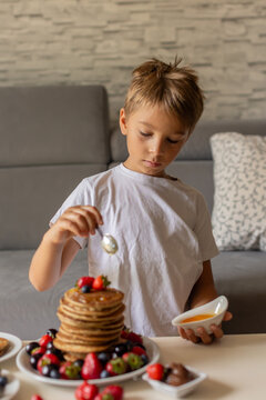 Sweet toddler child, boy, eating american pancakes with strawberries, cherries and blueberries, topped with syrup and drinking fresh orange juice