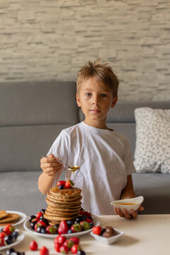 Sweet toddler child, boy, eating american pancakes with strawberries, cherries and blueberries, topped with syrup and drinking fresh orange juice