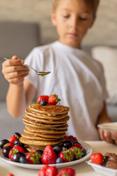 Sweet toddler child, boy, eating american pancakes with strawberries, cherries and blueberries, topped with syrup and drinking fresh orange juice