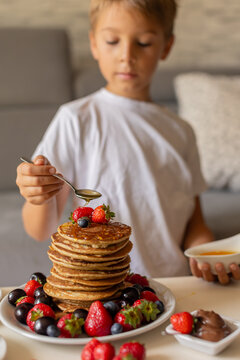Sweet toddler child, boy, eating american pancakes with strawberries, cherries and blueberries, topped with syrup and drinking fresh orange juice