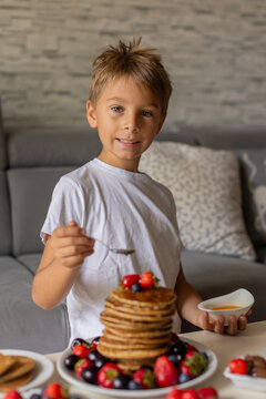 Sweet toddler child, boy, eating american pancakes with strawberries, cherries and blueberries, topped with syrup and drinking fresh orange juice