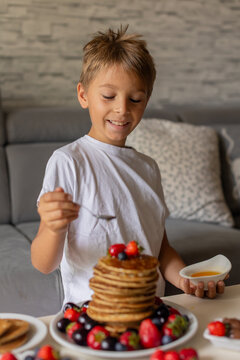 Sweet toddler child, boy, eating american pancakes with strawberries, cherries and blueberries, topped with syrup and drinking fresh orange juice