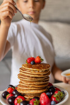 Sweet toddler child, boy, eating american pancakes with strawberries, cherries and blueberries, topped with syrup and drinking fresh orange juice