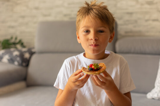 Sweet toddler child, boy, eating american pancakes with strawberries, cherries and blueberries, topped with syrup and drinking fresh orange juice