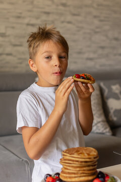 Sweet toddler child, boy, eating american pancakes with strawberries, cherries and blueberries, topped with syrup and drinking fresh orange juice