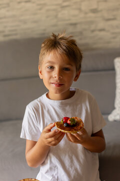 Sweet toddler child, boy, eating american pancakes with strawberries, cherries and blueberries, topped with syrup and drinking fresh orange juice
