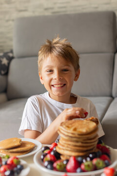Sweet toddler child, boy, eating american pancakes with strawberries, cherries and blueberries, topped with syrup and drinking fresh orange juice