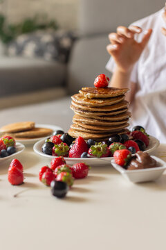 Sweet toddler child, boy, eating american pancakes with strawberries, cherries and blueberries, topped with syrup and drinking fresh orange juice