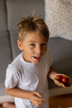 Sweet toddler child, boy, eating american pancakes with strawberries, cherries and blueberries, topped with syrup and drinking fresh orange juice