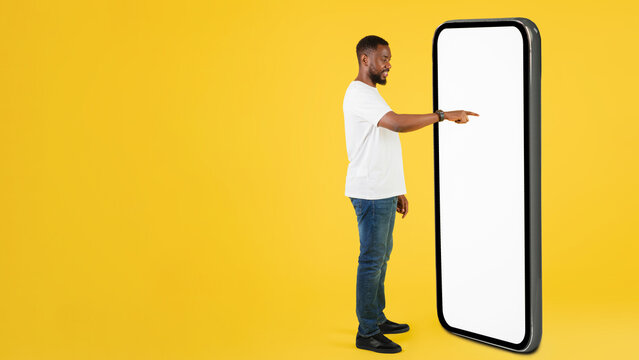 Focused Black Male Young Adult Pointing At Large Smartphone Display With Empty Screen In Studio Setting, Engaging With Technology. Business Concept.