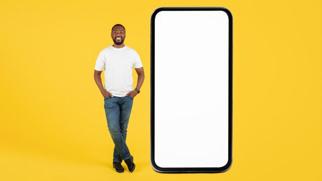 Smiling Black Man Standing With Hand In Pocket Next To Smartphone With Blank Screen In Studio. Ideal For Mockup And Advertising Use, Set Against A Bright Yellow Background, Engaging Concept.