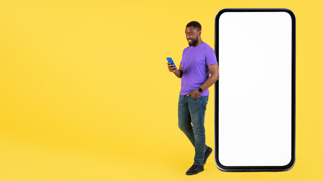 Smiling Black Man Enjoying His Smartphone, Standing Next To Large Mockup Phone On Bright Yellow Background. Engaged And Happy In Personal Moment, Showcasing Technology And Connectivity Concept.