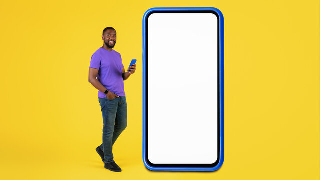 Smiling Black Male In Casual Attire Holding Smartphone With Empty Screen, Standing In Front Of Colorful Yellow Background, Showcasing Modern Technology And Mobile Advertisement Concept.
