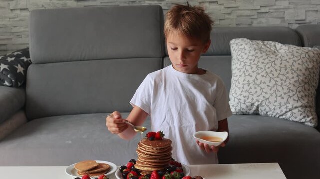 Sweet toddler child, boy, eating american pancakes with strawberries, cherries and blueberries, topped with syrup and drinking fresh orange juice