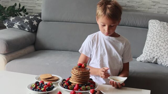 Sweet toddler child, boy, eating american pancakes with strawberries, cherries and blueberries, topped with syrup and drinking fresh orange juice