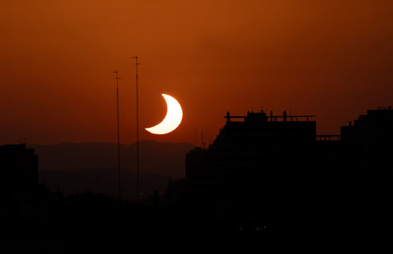 Partial solar eclipse crescent sun glows beside the dark silhouette of a tall building, thin antenna masts, orange sky, layered mountain ridge, city rooftops in shadow