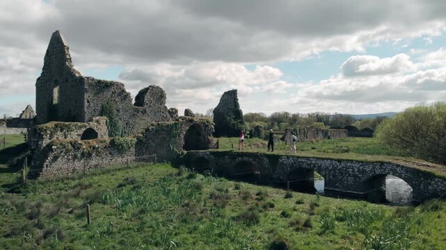 People walking across ancient stone bridge towards historic Athassel Abbey ruins in Tipperary, Ireland, exploring centuries-old architecture and lush green countryside, conveying a journey through tim