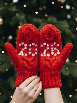 Red knitted mittens displaying 2023 with snowflakes against a blurred christmas tree background