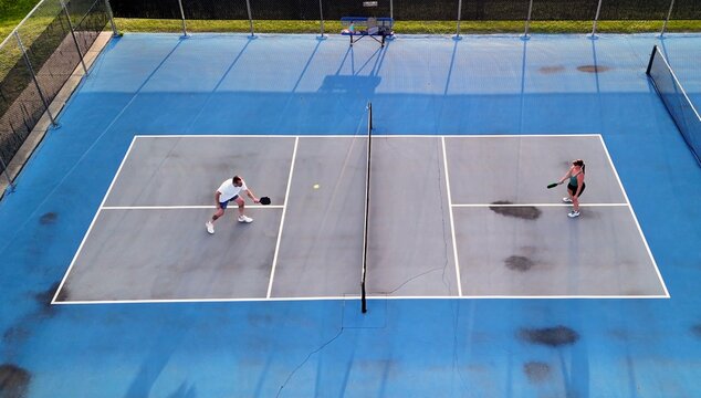Aerial overhead view of a man and woman playing competitive pickleball game on outdoor blue courts, serving, volleying, and tapping paddles at the net