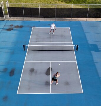 Aerial overhead view of a man and woman playing competitive pickleball game on outdoor blue courts, serving, volleying, and tapping paddles at the net