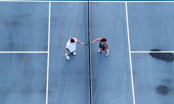 Aerial overhead view of a man and woman playing competitive pickleball game on outdoor blue courts, serving, volleying, and tapping paddles at the net
