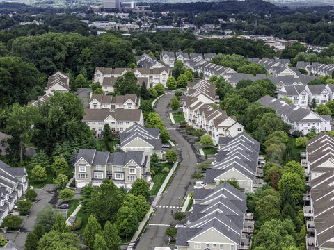 Aerial view of a suburban neighborhood featuring rows of residential houses with dark shingled roofs, lush green trees, and winding streets in Patterson, NY, United States.
