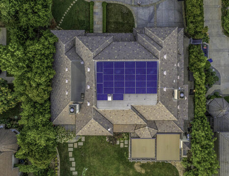 Aerial view of rooftop photovoltaic arrays and solar panels installed on a residential house surrounded by lush green trees and a landscaped backyard in Universal City, California, United States.