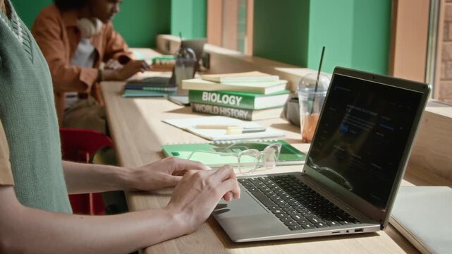 Unknown student typing on laptop in library with textbooks in background. Ideal for promoting online education platforms, student software discounts or university programs