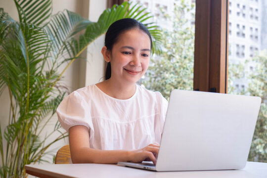 Smiling woman working on a laptop in a bright modern workspace with natural light, representing remote work, home office, online business, digital productivity, flexible lifestyle, and technology.