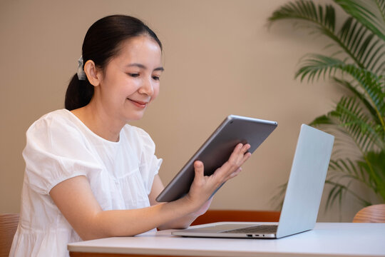 Smiling woman using a tablet and laptop in a modern workspace, representing remote work, digital productivity, online learning, multitasking, business technology, and flexible work lifestyle.