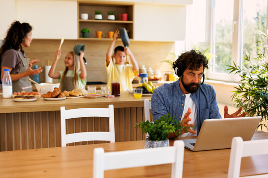 Overwhelmed father working from home at a laptop wearing headphones, while noisy children and mother are in the background