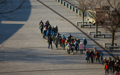 Group of school children in colorful winter clothes walking in a line across a paved city square. High-angle shot of a student field trip in an urban environment on a bright sunny day. © Kopylov.Design