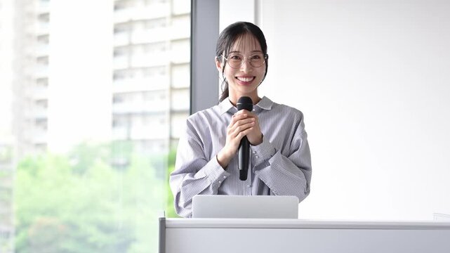 会議室でマイクを持ってプレゼンテーションを行う30代の日本人ビジネスパーソンの女性
